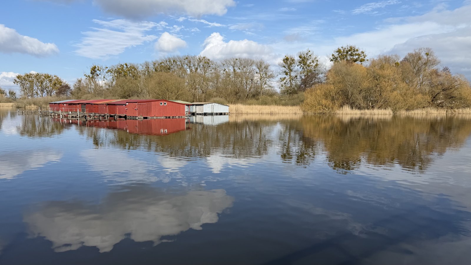 A calm lake at dusk with small red boathouses on the far shore, mirrored in the water.