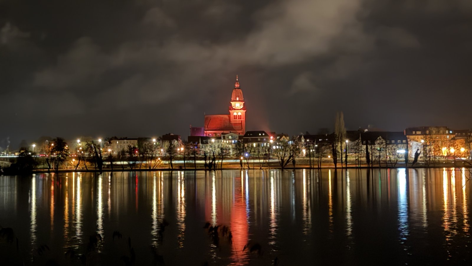 A small town at night with a brightly lit red church, reflected on dark water.