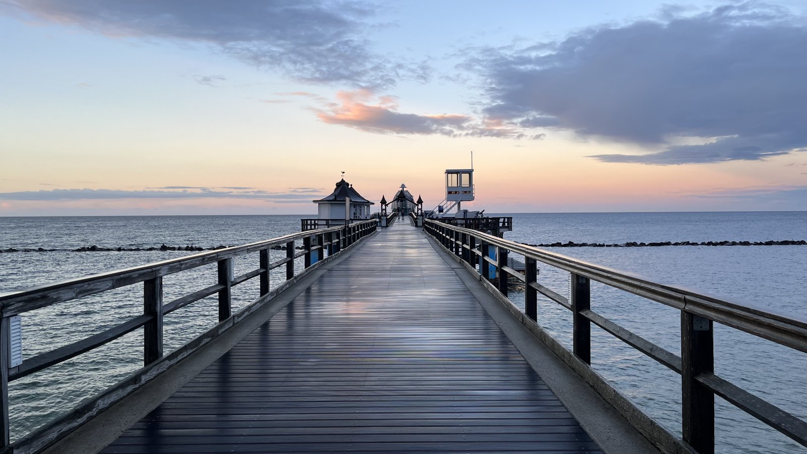 A long wooden pier with railings reaching out across calm water toward a distant shore.
